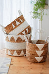 Three handcrafted banana leaf baskets with white geometric patterns and macrame details, placed on a wooden floor against a white background with a green plant.
