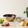 Two metallic bowls on a light surface with fruits and a plant.