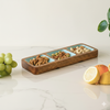 Wooden tray with compartments filled with nuts on a marble surface with fruits and a plant.
