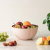 Pink fruit bowl filled with various fruits on a light surface with a plant in the background