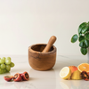 Wooden mortar and pestle on a marble surface with fruits and a plant in the background
