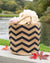 Woven tote bag with black zigzag pattern on a wooden surface near a body of water.
