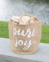 Woven basket with 'pure joy' text and a blanket on a wooden surface with a blurred natural background