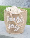 Woven basket with 'pure joy' text and a blanket on a grassy background