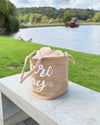Textile basket with 'Joy' printed on it, placed on a wooden surface by a lake.