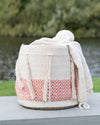 Woven bag with pink pattern and white tassels on a wooden surface with a blurred natural background