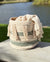 Decorative basket with tassels and a blanket on a wooden surface by a lake.