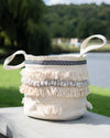 Decorative woven basket with fringe and handles on a wooden surface outdoors.