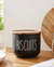 Black 'Biscuits' container on a wooden table with a jar of oats and a wooden board in the background.