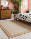 Beige braided rug in a bedroom with pink walls, wooden dresser, and bed with patterned blanket.