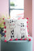 Decorative pillow with giraffe design and pink pom-poms on a teal stool against a pink wall.