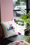 Decorative pillow with owl design on a window sill with plants and pink wall.