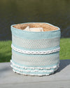 Woven basket with blue, white, and beige stripes on a wooden surface with grass and water in the background