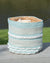 Woven basket with blue, white, and beige stripes on a wooden surface with grass and water in the background