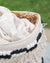 Woven basket with a white blanket and black and white beaded trim against a blurred green background