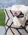 Decorative woven basket with tassels and a blanket draped over it, set against a natural background.