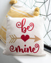 Decorative pillow with 'be mine' text and heart design on a surface.
