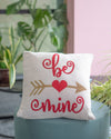 Decorative pillow with 'be mine' text on a chair with plants in the background