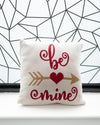 Decorative pillow with 'be mine' text and arrow design on a geometric patterned wall background