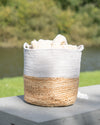 Woven basket with a natural base and white top, filled with stones, on a wooden surface with a blurred natural background.
