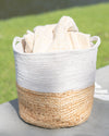 Woven basket with a white top and natural bottom, containing folded towels, on a surface with a grassy background.